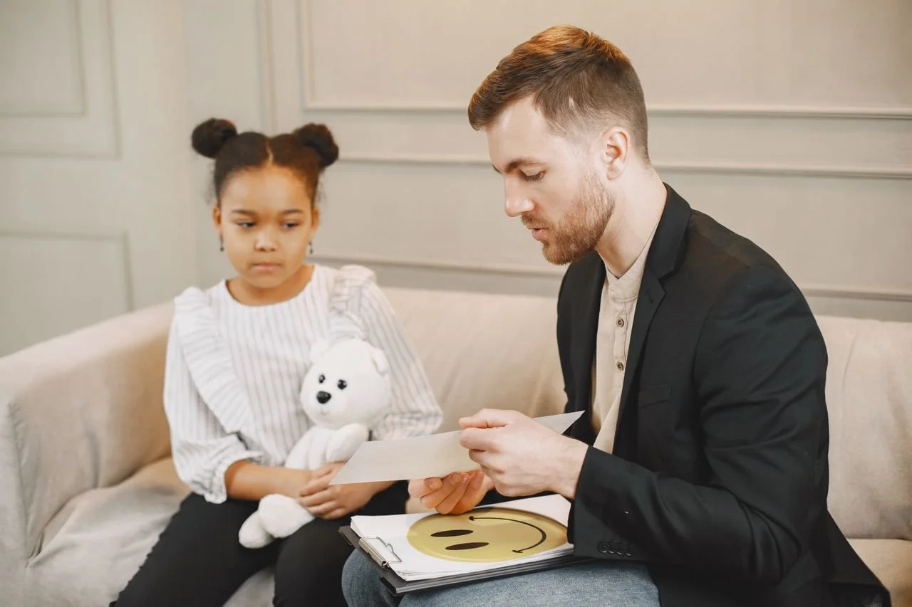about-01 A child holding a plush toy listens to a man in a therapy session.